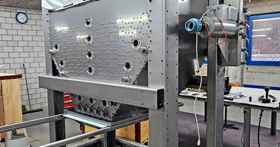 Das Bild zeigt eine große, rechteckige Metallkonstruktion in einer Werkstatt. Die Konstruktion besteht aus mehreren silbernen Platten, die mit vielen Schrauben verbunden sind. An der Vorderseite befinden sich mehrere runde Öffnungen. Rechts an der Seite ist ein Zylinder angebracht, der mit Kabeln und Rohren verbunden ist. Im Hintergrund sind Werkstattelemente wie eine Uhr an der Wand und ein Fenster zu sehen.