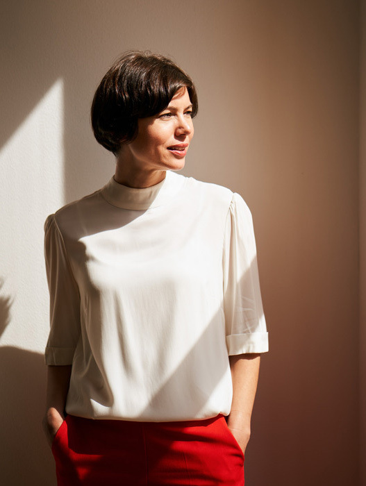 Eine junge Frau mit kurzen, dunklen Haaren blickt nach rechts in die Ferne. Sie trägt ein weißes Oberteil und eine rote Hose. Das Licht fällt von rechts auf sie und erzeugt Schatten an der Wand hinter ihr. Sie wirkt nachdenklich und ruhig.