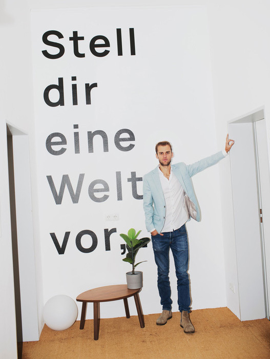 Ein Mann steht lächelnd vor einer weißen Wand mit dem Schriftzug "Stell dir eine Welt vor". Er trägt Jeans und eine hellblaue Jacke und deutet mit ausgestrecktem Arm auf eine offene Tür. Das Bild vermittelt eine positive und einladende Atmosphäre.