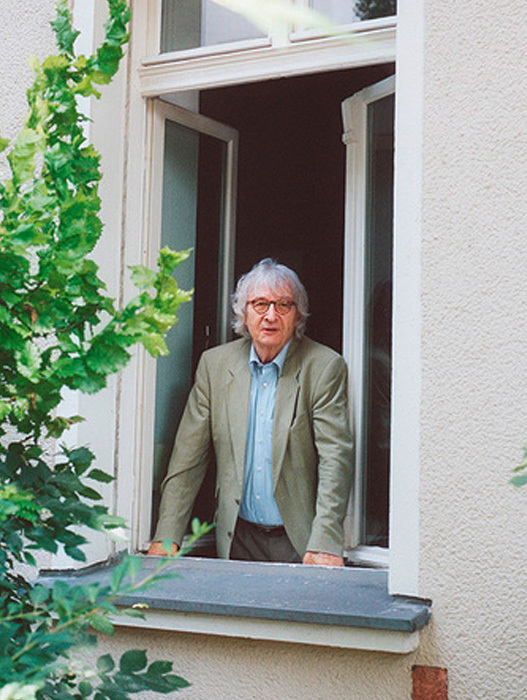 Ein älterer Mann mit weißem Haar lehnt lächelnd am Fensterbrett. Er trägt einen hellgrünen Blazer und ein blaues Hemd. Er scheint in eine angenehme Atmosphäre zu blicken. Das Fenster ist von grünen Pflanzen umrahmt und die Hauswand ist hell.