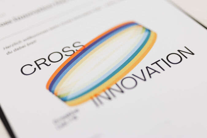 Das Bild zeigt eine Grafik auf einem weißen Blatt Papier. Die Grafik besteht aus einem farbigen, ovalen Ring, der in Regenbogenfarben verläuft. Darunter steht in großen, schwarzen Buchstaben das Wort "CROSS" und darunter das Wort "INNOVATION". Der Hintergrund ist unscharf und zeigt Textfragmente.