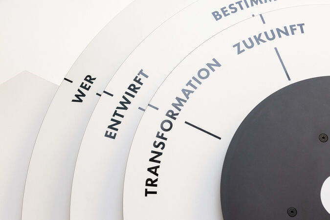 Das Bild zeigt eine Grafik in Form eines halbkreisförmigen Diagramms. Auf dem Diagramm sind die Begriffe "WER", "ENTWIRFT", "TRANSFORMATION" und "BESTIMMT ZUKUNFT" in einer spiralförmigen Anordnung angeordnet. Die Farben sind Weiß und Grau. Es vermittelt den Eindruck eines Prozesses oder einer Entwicklung.
