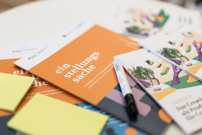 Mehrere orangefarbene Broschüren und Bücher liegen auf einem dunklen Untergrund. Die Broschüren sind mit Text versehen, der sich auf "Stellungsnahme" und "Fachkräfte" bezieht. Ein schwarzer Stift liegt darauf. Das Bild vermittelt einen Eindruck von Arbeit, Bildung oder kreativer Tätigkeit.