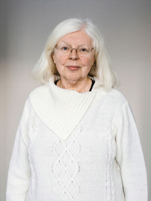 Porträt einer älteren Frau mit weißem Haar und Brille. Sie trägt einen weißen Pullover und blickt freundlich in die Kamera. Sie wirkt ruhig und gelassen.