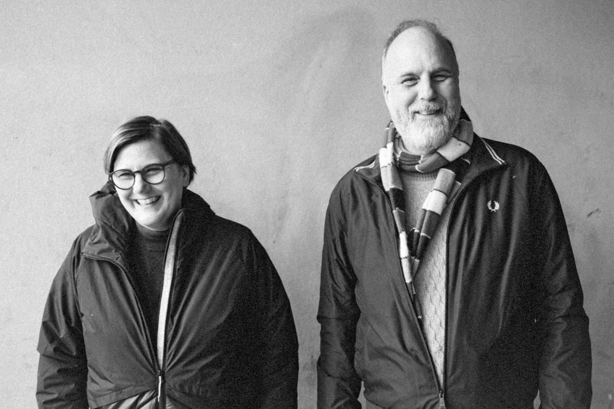 Schwarz-Weiß Porträt von zwei Personen. Links eine Frau mit Brille, die lächelt und eine dunkle Jacke trägt. Rechts ein Mann mit Bart, der ebenfalls lächelt und eine Jacke mit Schal trägt. Beide stehen vor einem hellen Hintergrund und wirken freundlich und entspannt.
