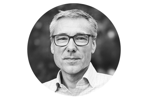 Schwarz-Weiß Porträt eines Mannes mittleren Alters mit kurzem, grau meliertem Haar und Brille. Er trägt ein kariertes Hemd und blickt freundlich in die Kamera. Er wirkt sympathisch und zugewandt.