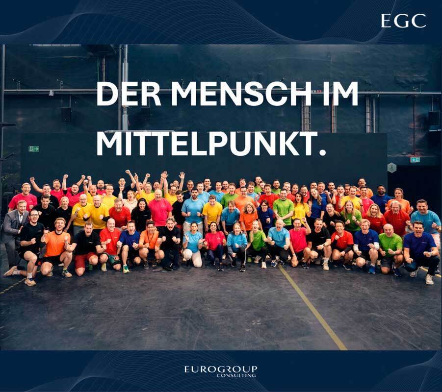 Das Bild ist eine Werbegrafik, die eine große Gruppe von Menschen zeigt, die gemeinsam vor einem dunklen Hintergrund posieren. In der Mitte steht der Text "DER MENSCH IM MITTELPUNKT.", was so viel bedeutet wie "Wir sind vereint in unserer Mission". Das Bild wirbt wahrscheinlich für ein Team oder eine Organisation, und die Verwendung eines dunklen Hintergrunds und des fettgedruckten Textes trägt dazu bei, die Aufmerksamkeit auf die Botschaft zu lenken.