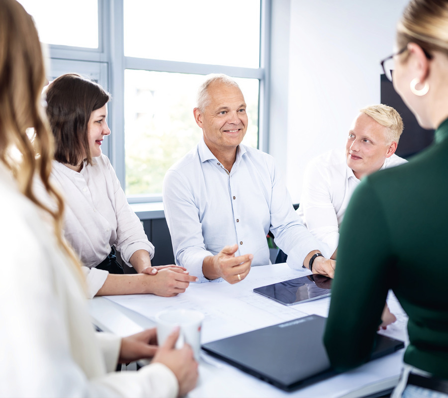 Das Bild zeigt eine Gruppe von Personen, die sich an einem Tisch treffen oder diskutieren. Es sind fünf Personen zu sehen: eine Person auf der linken Seite, zwei auf der rechten Seite und eine Person in der Mitte. Der Tisch ist mit einem weißen Tischtuch bedeckt, und es stehen zwei Laptops darauf, was auf eine gemeinschaftliche Arbeitsumgebung hindeutet. Die Personen scheinen auf das Gespräch konzentriert zu sein, wobei eine Person lächelt und eine andere in die Kamera blickt. Im Hintergrund sind ein Fenster, das den Blick nach draußen freigibt, und ein an der Wand montierter Fernsehbildschirm zu sehen.