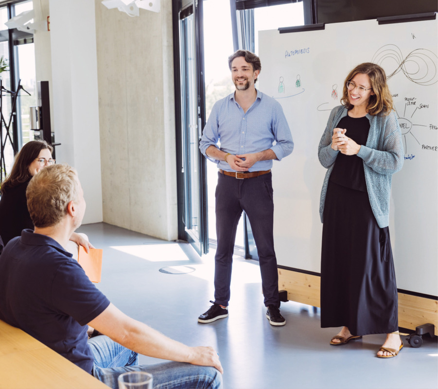Das Bild zeigt eine Gruppe von Personen in einem Raum mit einem Whiteboard und Diagrammen an der Wand. Ein Mann steht und präsentiert, während eine Frau steht und ihn anlächelt. Im Raum befinden sich weitere Personen, einige sitzen, andere stehen und sind in die Präsentation vertieft. Der Raum scheint gut beleuchtet zu sein, da natürliches Licht durch ein Fenster einfällt.