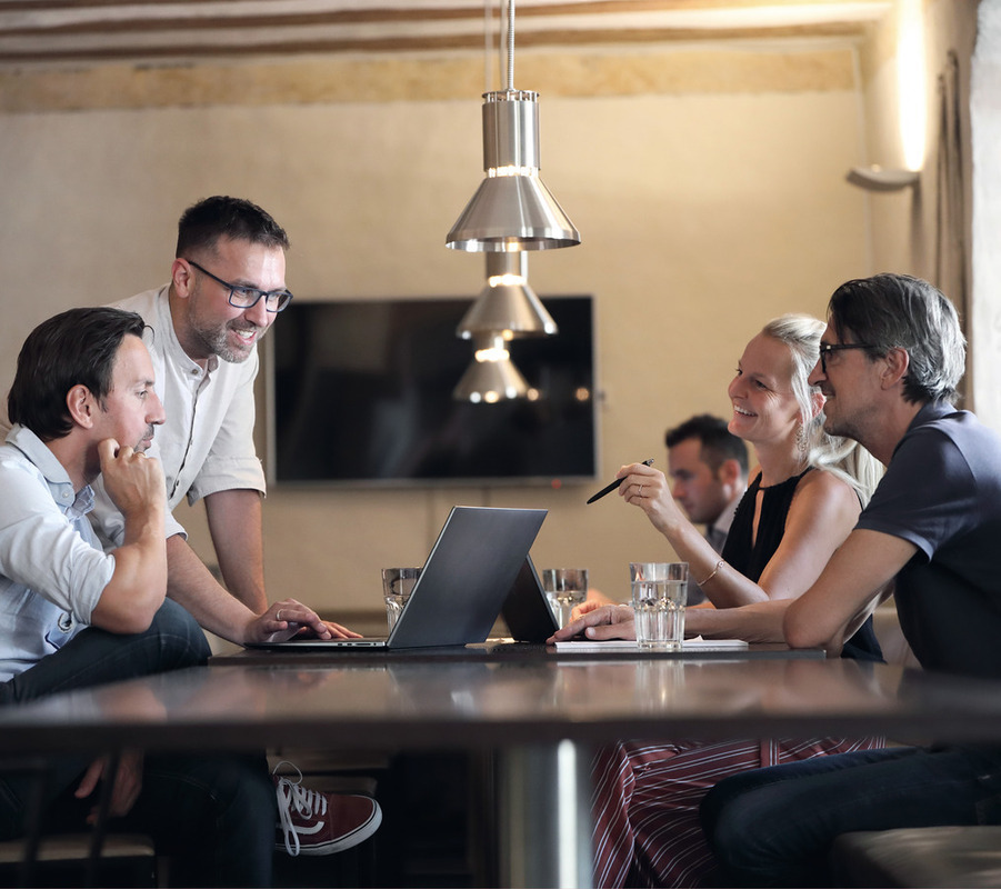 Eine Gruppe von Personen sitzt in einem Besprechungsraum um einen Tisch herum und diskutiert und arbeitet zusammen. Sie verwenden Laptops und andere Geräte, um gemeinsam zu arbeiten, während Tassen und Wasserflaschen auf dem Tisch stehen. Der Raum hat eine moderne und professionelle Atmosphäre, mit einem an der Wand montierten Fernseher und einer Pendelleuchte über dem Tisch.