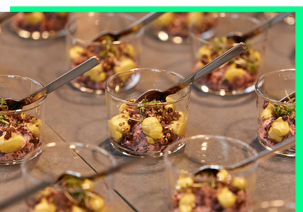 Das Bild zeigt eine Nahaufnahme von mehreren kleinen Glasbechern, die mit einem farbenfrohen und appetitlichen Lebensmittel gefüllt sind, möglicherweise einem Salat oder einem Dessert. Jeder Becher wird von einem Löffel begleitet, was darauf hindeutet, dass das Gericht mit einem Utensil genossen werden soll. Die Becher sind in einem gitterartigen Muster angeordnet, was eine geordnete und optisch ansprechende Präsentation ergibt. Der Hintergrund ist unscharf und lenkt die Aufmerksamkeit auf die Tassen und ihren Inhalt.