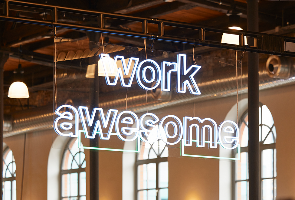 Das Bild zeigt ein Neonschild mit der Aufschrift "work awesome" in weißen Buchstaben. Das Schild hängt an einer Metallkonstruktion, wahrscheinlich einer Decke, und wird von einer Lichtquelle beleuchtet. Der Hintergrund des Bildes ist unscharf und lenkt die Aufmerksamkeit auf das Schild und seine Botschaft. Das Schild scheint Teil eines größeren Innenraums zu sein, möglicherweise ein Restaurant oder ein Büro, wobei im Hintergrund gewölbte Fenster zu sehen sind.