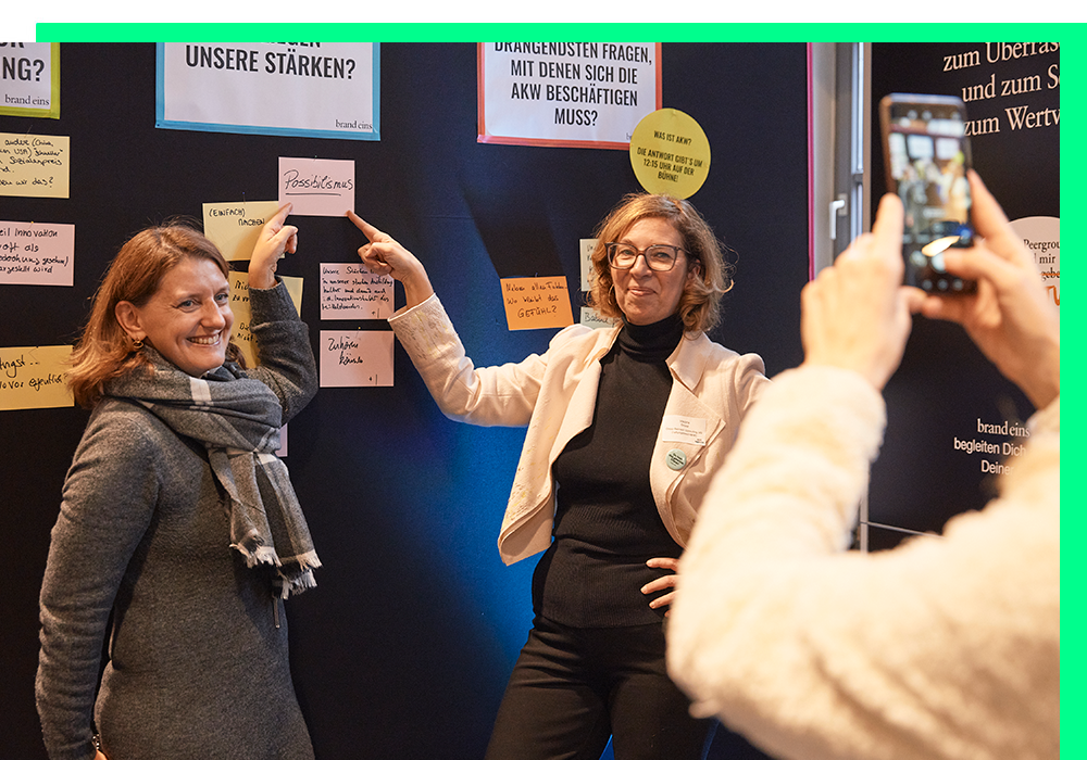 Das Bild zeigt zwei Frauen, die vor einem schwarzen Hintergrund stehen, an dem verschiedene Papiere und Notizen befestigt sind. Eine Frau hält ein Stück Papier hoch, während die andere mit einem Smartphone ein Foto von der Szene macht. Das Bild scheint eine Collage oder eine Präsentation mit mehreren Elementen zu sein, und der Schwerpunkt liegt auf der Interaktion zwischen den beiden Frauen und dem Akt des Fotografierens.