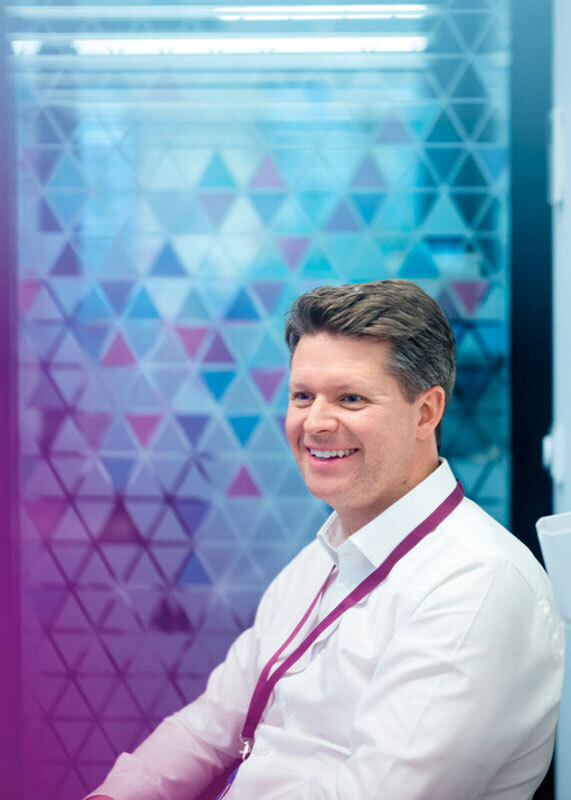 Ein Mann mit weißem Hemd und lila Schlüsselband lächelt in die Kamera, hinter ihm eine bunte, geometrisch gemusterte Wand.