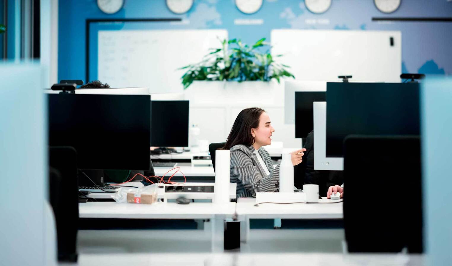 Das Bild zeigt eine Frau, die an einem Schreibtisch in einem Büro sitzt und an einem Computer arbeitet. Sie trägt eine graue Jacke und scheint auf ihre Arbeit konzentriert zu sein. Das Büro hat eine moderne und professionelle Atmosphäre, mit mehreren Schreibtischen, Computermonitoren und einer Topfpflanze im Hintergrund. Das Gesamtlayout und die Gestaltung des Raums lassen auf einen gut organisierten und funktionalen Arbeitsbereich schließen.