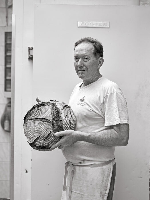 Eine Schwarz-Weiß-Fotografie eines Mannes, der vor einer Tür steht und einen großen Laib Brot hält. Der Mann trägt ein weißes Hemd und eine Schürze, und über der Tür steht ein Schild mit der Aufschrift "Foster".