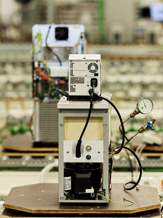 Das Bild zeigt eine Nahaufnahme eines komplexen elektronischen Geräts, an dem verschiedene Komponenten und Drähte angebracht sind. Das Gerät scheint ein kleiner, weißer Kasten zu sein, an dem ein Bedienfeld und ein Manometer angebracht sind. Das Gerät steht auf einer hölzernen Oberfläche, und der unscharfe Hintergrund lässt auf eine Industrie- oder Laborumgebung schließen. Das Bild enthält keinen erkennbaren Text, und die Position des Geräts und der unscharfe Hintergrund deuten darauf hin, dass es der Hauptfokus des Bildes ist.