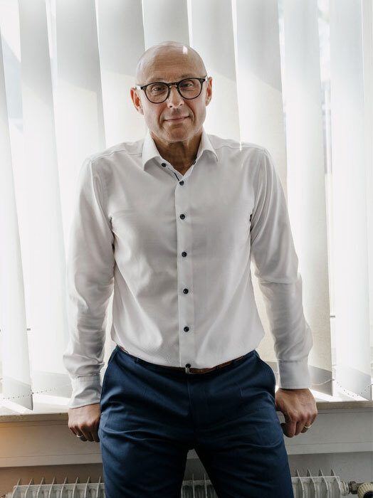 Das Bild zeigt einen Mann, der vor einem Fenster mit vertikalen Jalousien steht. Er trägt ein weißes Hemd, eine blaue Hose und eine Brille. Der Mann lächelt und scheint für die Kamera zu posieren.