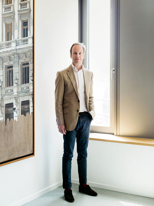 Ein Mann in einem beigen Blazer und einer dunklen Hose steht in einem Raum mit einem großen Fenster und lehnt an der Wand. Hinter ihm ist ein Gemälde eines Gebäudes an der Wand zu sehen.