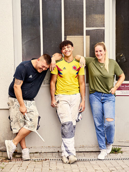 Drei Freunde stehen zusammen an einer Wand, lächeln und posieren für ein Foto. Sie sind leger gekleidet, wobei eine Person ein gelbes Hemd und die beiden anderen ein grünes und blaues Hemd tragen. Im Hintergrund sind ein Fenster und ein Schild zu sehen, die den Kontext der Szene verdeutlichen.