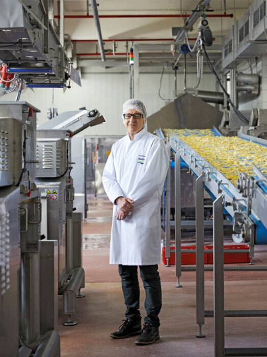 Ein Mann in einem weißen Laborkittel steht in einer großen Industrieküche, umgeben von verschiedenen Maschinen und Geräten. Der Mann scheint ein Koch oder Lebensmittelverarbeiter zu sein, da er einen weißen Kittel und ein schützendes Haarnetz trägt. Die Küche ist mit verschiedenen Maschinen und Geräten gefüllt, darunter ein Förderband mit gelben Gegenständen, die auf eine Lebensmittelproduktion oder -verarbeitung hindeuten.