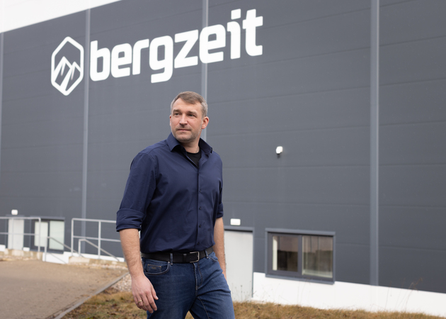 Das Bild zeigt einen Mann, der vor einem großen Industriegebäude mit einem auffälligen Logo steht. Der Mann ist mit einem blauen Hemd und Jeans bekleidet und scheint zur Seite zu schauen. Das Gebäude hat ein graues Äußeres, und das Logo ist gut sichtbar an der Seite des Gebäudes angebracht.