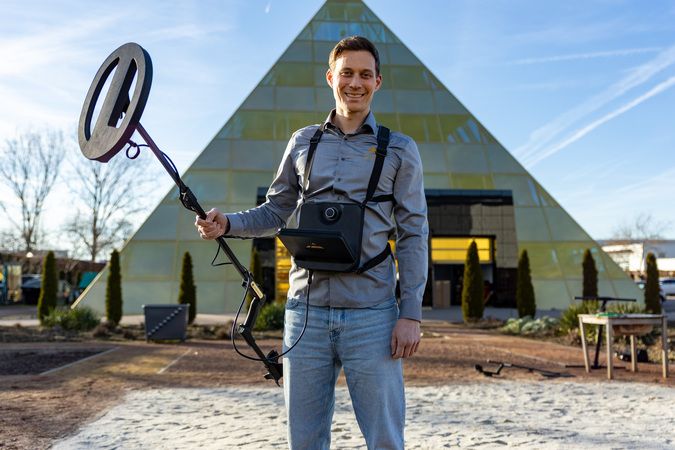 Ein Mann steht vor einem großen, modernen Gebäude mit einer dreieckigen Form und hält einen Metalldetektor in der Hand. Er trägt ein graues Hemd und eine blaue Jeans, und er scheint zu lächeln. Das Bild wird als für Menschen mit Behinderungen zugänglich beschrieben und entspricht den Richtlinien des EAA (Emergency Alert and Alert System).