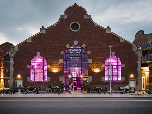 Das Bild zeigt ein großes Backsteingebäude mit gewölbten Fenstern und einer markanten Kuppel, die von violetten Lichtern beleuchtet wird. Vor dem Gebäude sitzen mehrere Personen, die möglicherweise ein gesellschaftliches Beisammensein oder eine Veranstaltung genießen. Die Szene spielt sich vor einem bewölkten Himmel ab, der eine stimmungsvolle Atmosphäre schafft. Das Bild ist in Farbe, wobei sich die violetten Lichter von der Backsteinfassade abheben.