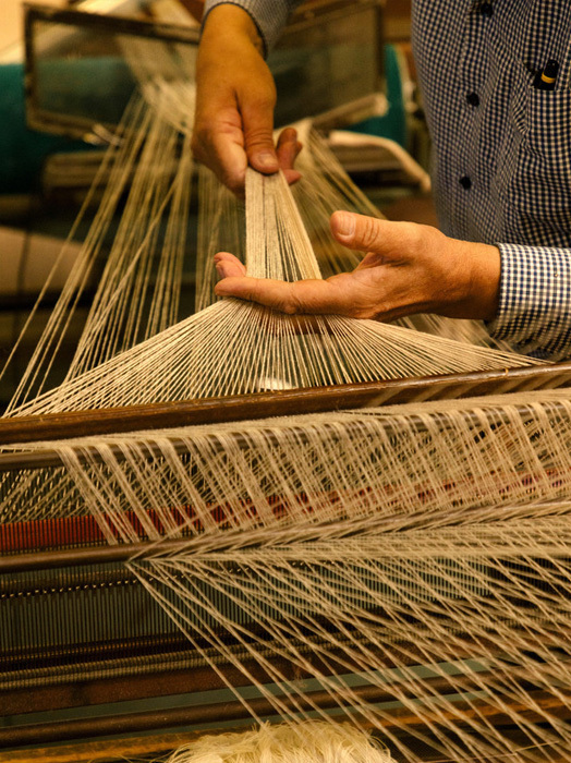 Das Bild zeigt eine Nahaufnahme der Hände einer Person, die an einem Webstuhl mit zahlreichen Fäden arbeitet. Die Person hält die Fäden in der Hand und scheint dabei zu sein, sie zu weben oder zu stricken. Das Bild konzentriert sich auf die komplizierten Details des Webstuhls und die Handwerkskunst, die mit der Herstellung des Gewebes verbunden ist.