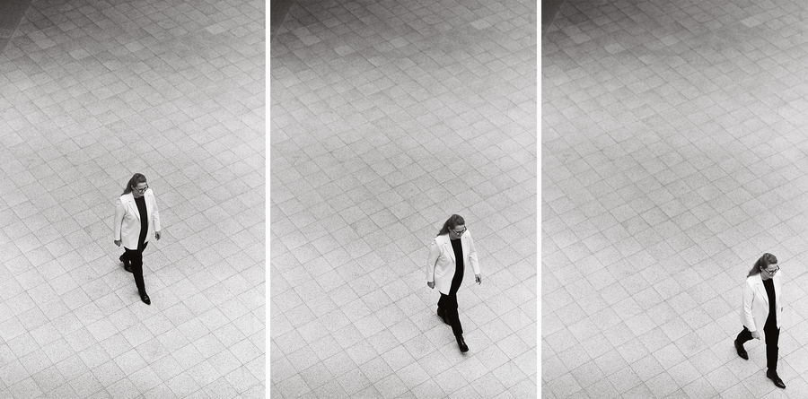 Bei dem Bild handelt es sich um eine Schwarz-Weiß-Collage aus drei Fotografien, die eine Frau zeigen, die auf einem gefliesten Boden geht. Die Frau trägt einen weißen Mantel und eine schwarze Hose, und ihre Position und Bewegung sind in verschiedenen Bildern festgehalten. Die Collage ist so gestaltet, dass sie für ein breites Publikum, einschließlich sehbehinderter Personen, zugänglich ist, da sie eine klare und übersichtliche Darstellung des Themas bietet.