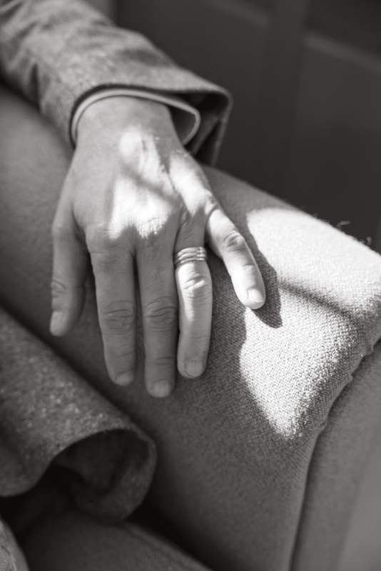 Bei dem Bild handelt es sich um eine Schwarz-Weiß-Fotografie der Hand einer Person, die auf einem Sofa ruht. Die Hand trägt einen Ehering, und die Person scheint eine entspannte Haltung einzunehmen, wobei die Finger sanft um die Armlehne des Sofas geschlungen sind. Der Hintergrund ist unscharf und lenkt den Fokus auf die Hand und das Sofa.