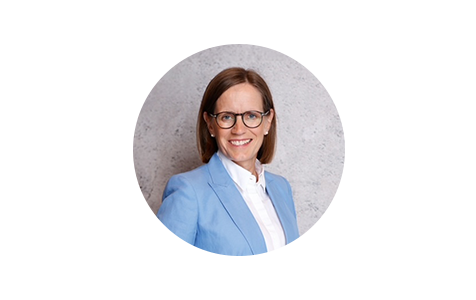 Das Bild zeigt eine Frau, die einen blauen Blazer und eine Brille trägt und in die Kamera lächelt. Sie steht vor einem grauen Hintergrund, der eine Wand zu sein scheint. Das Bild ist quadratisch und enthält keinen Text oder zusätzliche Objekte. Die Pose und die Kleidung der Frau lassen auf einen professionellen oder formellen Rahmen schließen, aber der spezifische Kontext oder Zweck des Bildes ist nicht klar.