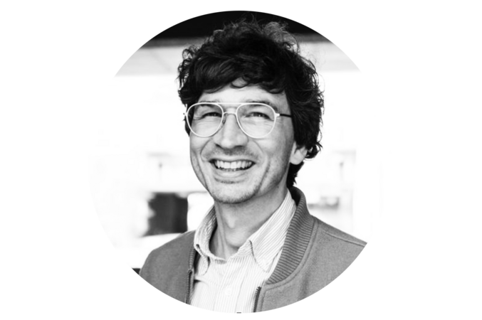 Das Bild ist ein Schwarz-Weiß-Porträt eines lächelnden Mannes, der eine Brille und eine Jacke trägt. Der Mann blickt mit einem leichten Lächeln zur Seite. Der Hintergrund ist unscharf und lenkt die Aufmerksamkeit auf den Mann und seinen Ausdruck. Das Bild enthält keinen erkennbaren Text, und die Position des Mannes und die Hintergrundelemente suggerieren eine Konzentration auf das Motiv.