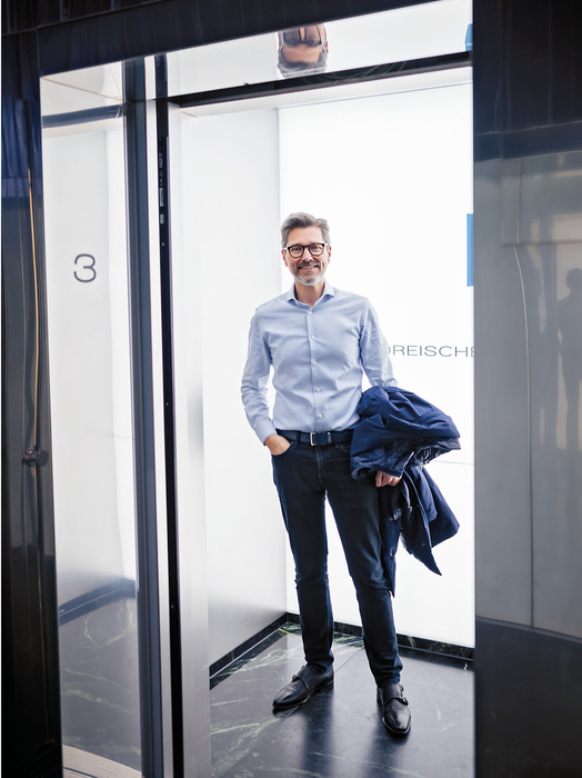 Ein Mann mit blauem Hemd und Brille steht in einem Aufzug, lächelt und hält eine Jacke. Der Aufzug hat eine Glastür und die Nummer 3 ist an der Wand zu sehen.