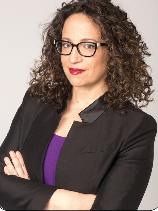 Das Bild zeigt eine Frau mit lockigem Haar, die eine Brille und einen schwarzen Blazer trägt. Sie steht mit verschränkten Armen und schaut mit neutralem Blick direkt in die Kamera. Der Hintergrund ist schlicht, was dazu beiträgt, die Aufmerksamkeit auf die Frau und ihre Kleidung zu lenken.