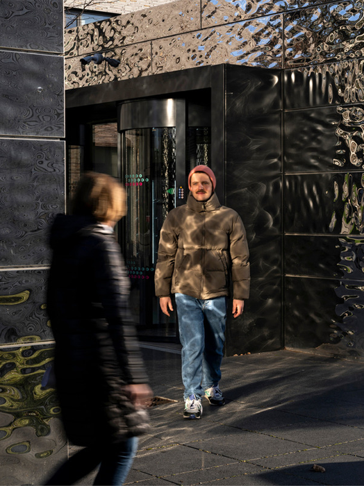 Das Bild zeigt einen Mann und eine Frau, die auf einem Gehweg vor einem Gebäude mit einer schwarz-grauen Fassade gehen. Der Mann trägt eine braune Jacke und eine rote Mütze, während die Frau einen schwarzen Mantel trägt. Sie scheinen sich zu unterhalten oder einfach die Gesellschaft des anderen zu genießen. Das Bild wurde aus einem niedrigen Winkel aufgenommen, wodurch der Mann und die Frau größer und deutlicher im Bild erscheinen.