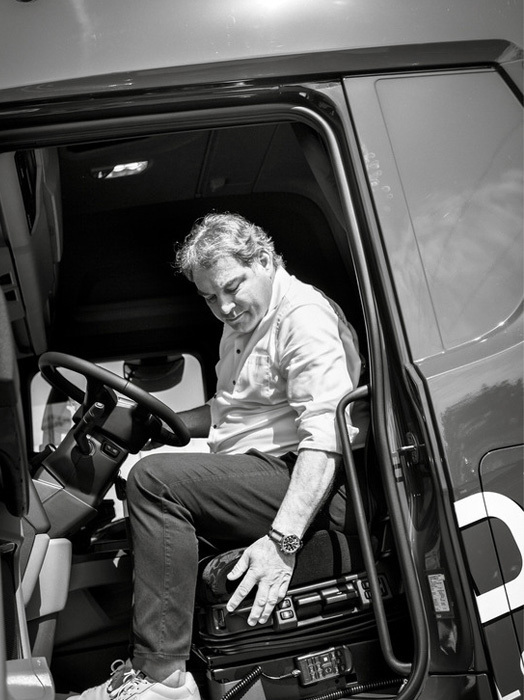 Das Bild ist eine Schwarz-Weiß-Fotografie eines Mannes, der auf dem Fahrersitz eines Lastwagens sitzt und auf das Lenkrad blickt. Der Lkw ist geparkt, und der Mann scheint sich entweder auf die Fahrt vorzubereiten oder gerade an seinem Ziel angekommen zu sein. Das Foto ist in Schwarz-Weiß gehalten, was ihm eine klassische und zeitlose Ausstrahlung verleiht. Die Tür des Lastwagens ist geöffnet, und der Mann sitzt auf dem Fahrersitz, der sich in der Mitte des Bildes befindet.