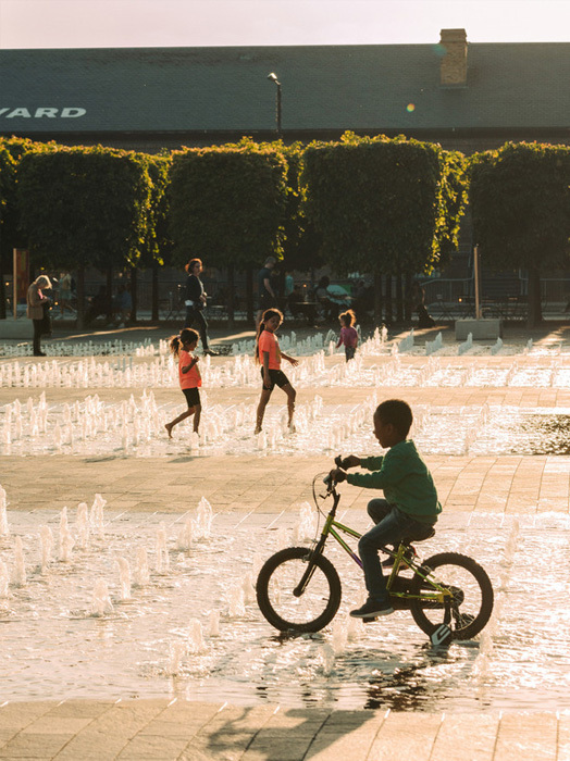 Das Bild zeigt eine lebhafte Szene in einem Park mit einem kleinen Jungen, der in der Nähe eines Springbrunnens Fahrrad fährt. Mehrere andere Kinder spielen im Wasser und schaffen eine fröhliche Atmosphäre. Im Hintergrund ist ein Gebäude mit dem Wort "YARD" zu sehen, was darauf hindeutet, dass sich der Park in einem Wohngebiet befindet. Das Bild ist aus einem niedrigen Winkel aufgenommen, wodurch der Junge auf dem Fahrrad und die im Wasser spielenden Kinder hervorgehoben werden.