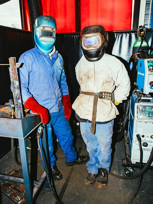 Das Bild zeigt zwei Personen mit Schutzausrüstung, einschließlich Helmen und Handschuhen, die in einer Werkstattumgebung stehen. Sie scheinen an einer Maschine oder Ausrüstung zu arbeiten, die im Hintergrund zu sehen ist. Die Szene ist vor einem roten Vorhang dargestellt, der einen kontrastreichen Hintergrund für die industrielle Umgebung bildet. Das Bild ist in Farbe, und die beiden Personen stehen nahe beieinander, was auf eine kollaborative oder teamorientierte Atmosphäre hindeutet.