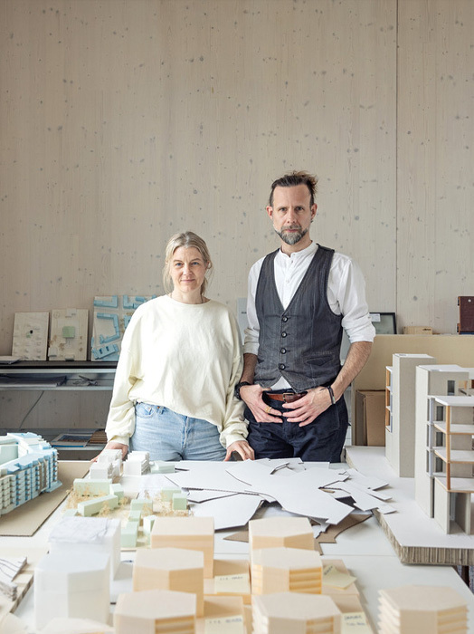 Auf dem Bild stehen ein Mann und eine Frau vor einem Tisch, der mit Architekturmodellen und Skizzen gefüllt ist. Der Mann trägt eine Weste, während die Frau einen weißen Pullover anhat. Der Tisch ist mit verschiedenen architektonischen Modellen und Papieren bedeckt, was auf einen Arbeitsbereich für architektonische Entwürfe oder Planungen hindeutet.