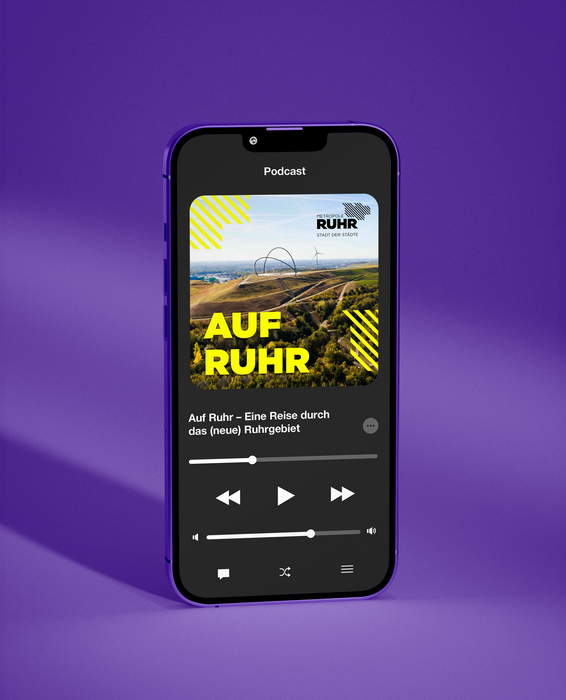 Das Bild zeigt ein lilafarbenes Smartphone, das eine Podcast-Oberfläche mit einer malerischen Landschaft auf dem Bildschirm anzeigt. Der Podcast scheint den Titel "Auf Ruhr - Eine Reise durch das Neue Ruhrgebiet" zu tragen und wird gerade abgespielt. Das Telefon liegt auf einer lilafarbenen Oberfläche, und die Benutzeroberfläche umfasst eine Wiedergabetaste, einen Lautstärkeregler und eine Navigationsleiste. Das Gesamtdesign und das Layout suggerieren ein zugängliches und benutzerfreundliches Erlebnis für die Podcast-Hörer.