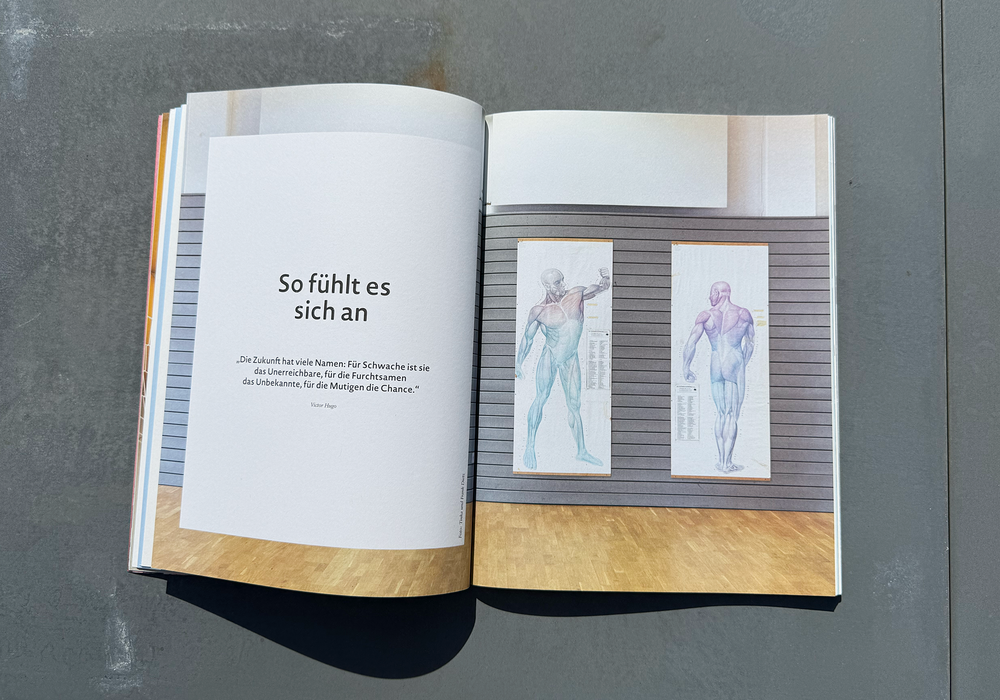 Das Bild zeigt ein aufgeschlagenes Buch mit Text und zwei Abbildungen von menschlichen Figuren auf der rechten Seite vor einem grauen Hintergrund.