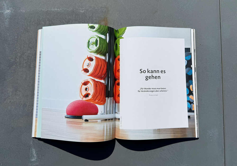 Das Bild zeigt ein aufgeschlagenes Buch mit einem Foto auf der linken Seite, das eine Sammlung bunter Trainingsgeräte, darunter Hanteln und Gewichtsscheiben, in einem Regal zeigt. Die rechte Seite enthält ein deutsches Zitat mit der Aufschrift "So kann es gehen", was übersetzt bedeutet: "Nicht die Übung ist wichtig, sondern der Geist." Das Buch scheint für eine zugängliche Website konzipiert zu sein, die sich an ein deutschsprachiges Publikum wendet.