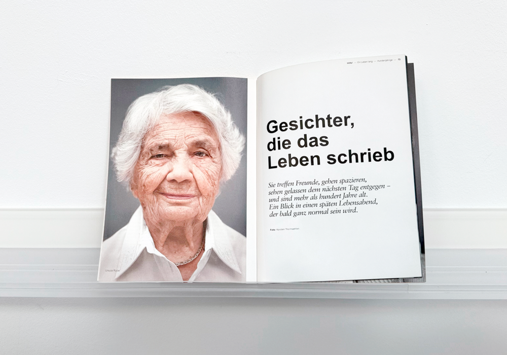 The image displays an open book with a photograph of an elderly woman on the left page and a German text on the right page. The text is in German and reads "Gesichter, die das Leben schrieb. So teine Frühen, gefänzen, sehen Frühen des nicht zum Tag entgen - und ande mühnde Tag entgen - Ein blick en ein spätlesend Lebensabend, der ganze normale seim word." The book is placed on a white shelf, and the image is in color.