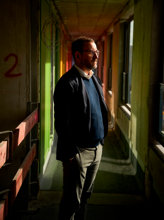 Das Bild zeigt einen Mann, der in einem Flur mit einer bunten Wand auf der einen Seite und einem Fenster auf der anderen Seite steht. Der Mann trägt eine dunkle Jacke, eine Brille und ein blaues Hemd. Er blickt zur Seite und beobachtet möglicherweise etwas oder jemanden außerhalb des Bildes. Der Flur scheint schwach beleuchtet zu sein, wobei durch das Fenster auf der rechten Seite Sonnenlicht einfällt, das einen Kontrast zwischen den hellen und dunklen Bereichen schafft.
