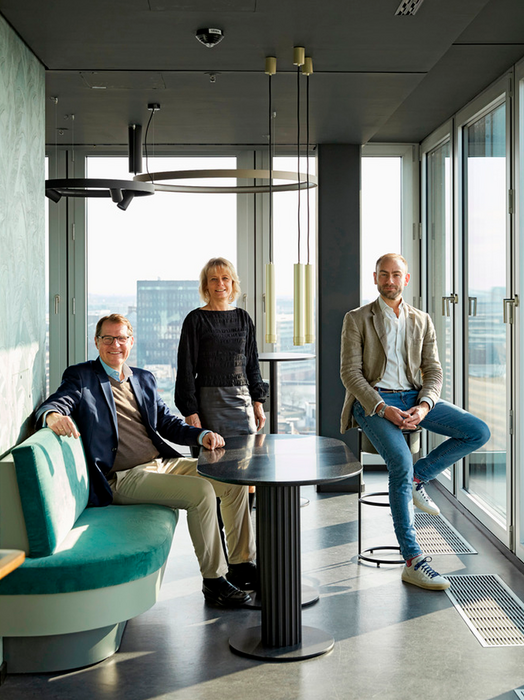 Drei Personen sitzen in einem modernen Büroraum mit großen Fenstern und Blick auf eine Stadtlandschaft. Der Raum ist mit einer runden, von der Decke hängenden Leuchte, einem schwarzen Tisch in der Mitte und einer grünen Bank auf der linken Seite ausgestattet. Die Personen sind geschäftsmäßig gekleidet. Eine Person trägt eine blaue Jacke und eine Brille, eine andere einen schwarzen Blazer und die dritte eine beigefarbene Jacke.