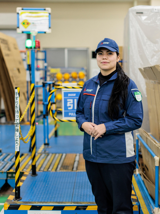 Das Bild zeigt eine Frau in einer Industrieumgebung, die eine blaue Uniform und eine Mütze trägt. Sie steht vor einer blau-gelb gestreiften Absperrung, bei der es sich offenbar um eine Sicherheitsmaßnahme handelt. Im Hintergrund sind verschiedene Geräte und Maschinen zu sehen, die auf eine Produktions- oder Fließbandumgebung hindeuten. Die Frau ist das Hauptmotiv des Bildes, und es gibt keine anderen erkennbaren Objekte oder Handlungen. Das Bild enthält keinen Text, und die relativen Positionen der Objekte und der Frau lassen auf eine typische Industrieszene schließen.