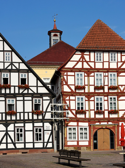 Das Bild zeigt einen bezaubernden europäischen Stadtplatz mit traditionellen Fachwerkhäusern in Weiß und Rot, die kunstvoll verziert und mit Blumenkästen versehen sind. Im Hintergrund erhebt sich ein Uhrenturm, der die malerische Szenerie noch unterstreicht. Im Vordergrund steht eine Bank, die die Passanten einlädt, sich zu setzen und die Aussicht zu genießen. Die gesamte Szene strahlt ein Gefühl von Geschichte und architektonischer Schönheit aus, wie es für eine europäische Stadt charakteristisch ist.