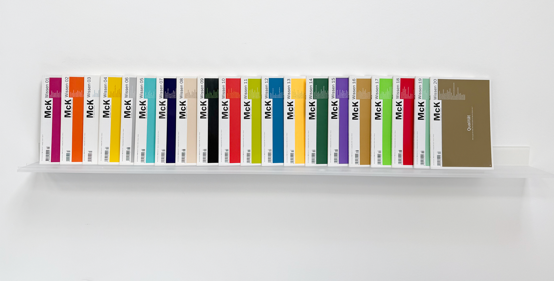 Das Bild zeigt eine Sammlung von bunten Büchern oder Zeitschriften, die ordentlich auf einem weißen Regal vor einem weißen Hintergrund angeordnet sind.