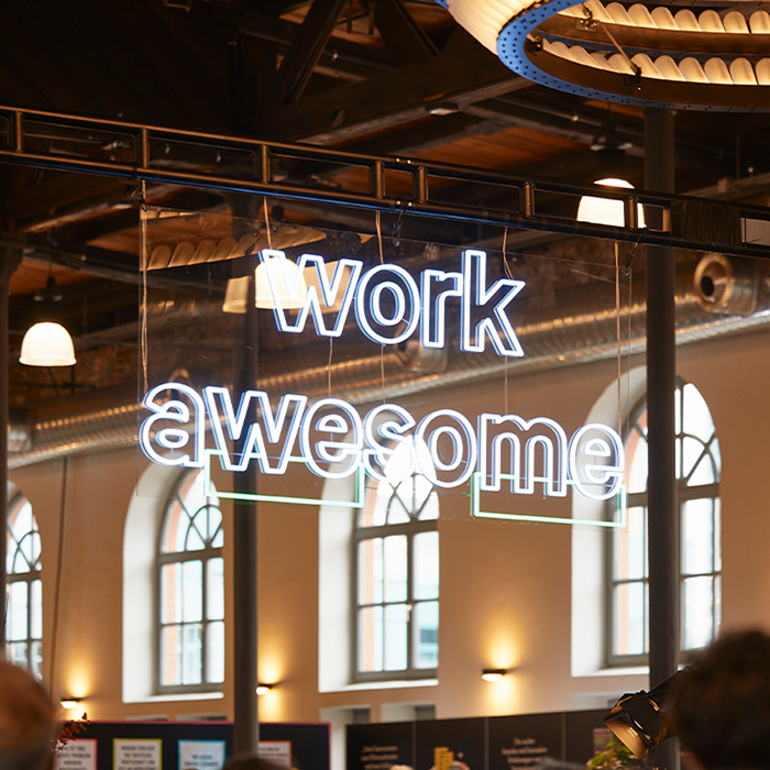 Das Bild zeigt ein Neonschild mit den Worten "work awesome" in einer modernen, serifenlosen Schrift. Das Schild ist von der Decke abgehängt und bildet den Hauptschwerpunkt des Bildes. Der Hintergrund des Bildes ist unscharf und deutet auf eine Innenumgebung hin, möglicherweise ein Restaurant oder ein Café. Im Hintergrund sind auch einige Personen zu sehen, die jedoch nicht im Mittelpunkt des Bildes stehen. Das Bild enthält keinen Text, der nicht deutlich sichtbar oder lesbar ist.